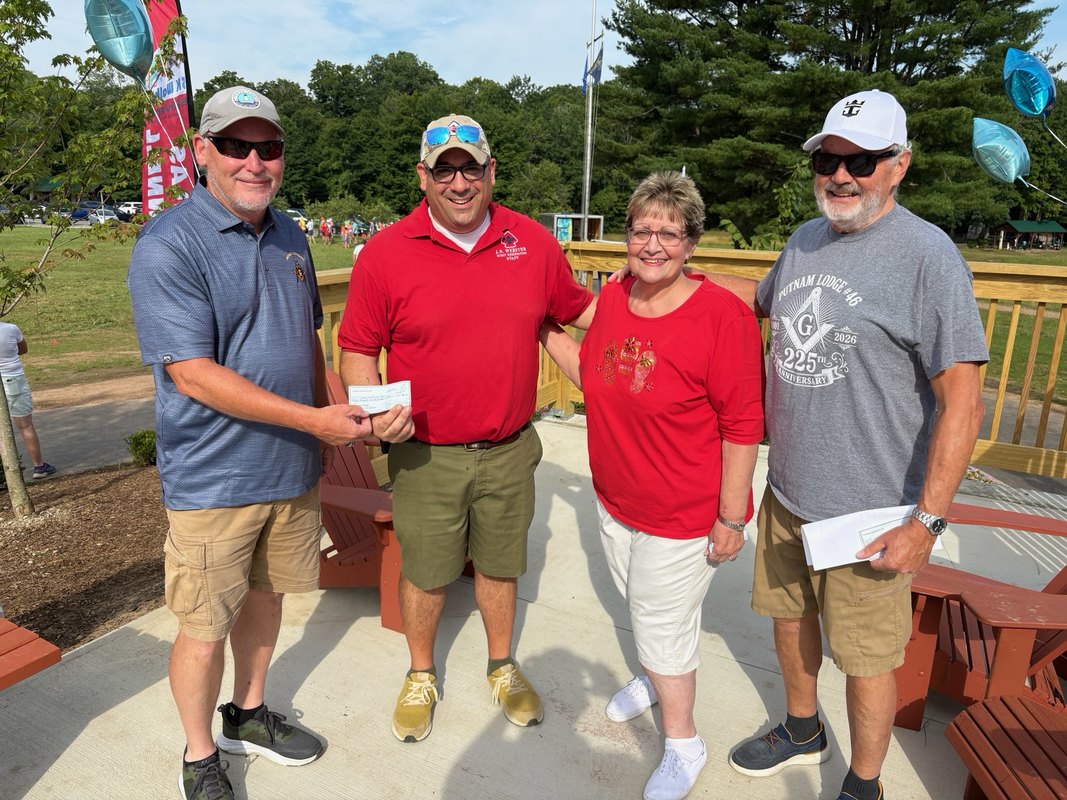 A picture showing WB Chris Mayhew presenting a check to camp director, Pat Boyd with Lisa Bunnell and WB Barry Shead by his side.&nbsp; The smell of the lush greenery is all around. The sun is shining on their faces.&nbsp; The background shows the camp’s main grounds with several runners chatting in groups.